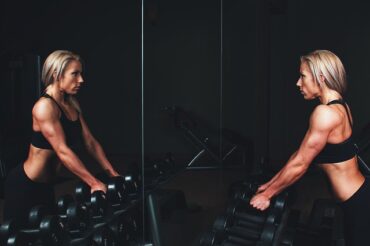 blonde haired woman looking at herself in a mirror. She has big muscles and is about to do some wieght lifting as there are a selection of hand weights in front of her. She likes to look good at the gym.