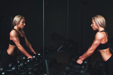 blonde haired woman looking at herself in a mirror. She has big muscles and is about to do some wieght lifting as there are a selection of hand weights in front of her. She likes to look good at the gym.