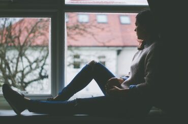 woman sat in a window looking out. the image is dull colours, very depressing and it suggests she is suffering with post natal anxiety