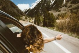 Brunette woman leaning out of the front of the car stretching her arm out. She is driving along a highway through the mountains and it is a beautiful sunny day. She is enjoying travel as a means of self care and reflection.