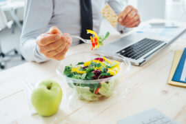 Close up of a bowl of salad on a` desk nexy to a computer kepboard and an apple. There is an male office workers torso and arms in shot, he is hokding a forful of salad in his left hand.