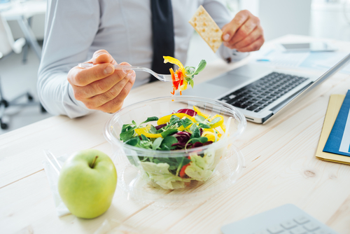 Close up of a bowl of salad on a` desk nexy to a computer kepboard and an apple. There is an male office workers torso and arms in shot, he is hokding a forful of salad in his left hand.