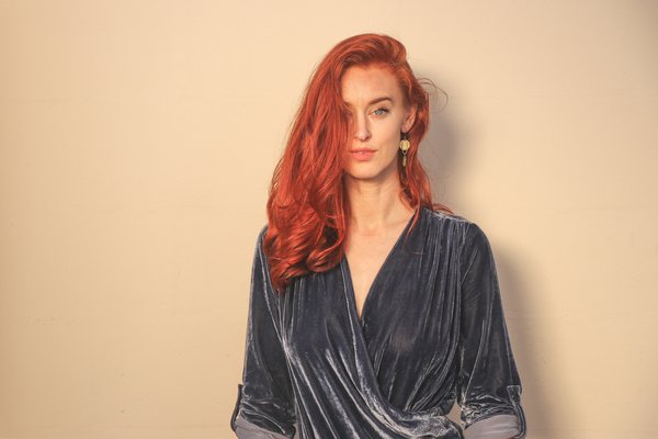 Long haired red headed woman standing against a plain neautrl background, wearing a mid grey velvet v necked long sleeved top.