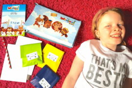 a young blonde haired girl lying on a pink rug next to the contents of the Weekend Box Club craft kits she has just received through the post