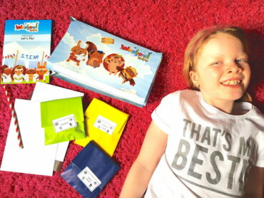 a young blonde haired girl lying on a pink rug next to the contents of the Weekend Box Club craft kits she has just received through the post