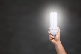 A black background with a persons bare arm holding up an energy light bulb on the right hand side of the picture