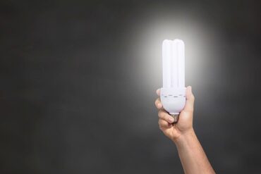 A black background with a persons bare arm holding up an energy light bulb on the right hand side of the picture