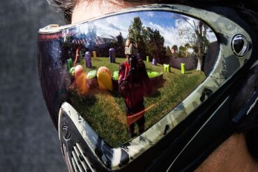 Close up of a pair of paintballing goggles on a mans face. Reflected in them is a paintballer aiming the gun and there are obstacles on the paintballing course in the background