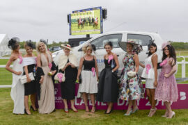 9 ladies dressed up for the horse races stood in front of a car and there is a TV screen with horces racing on it in the background.