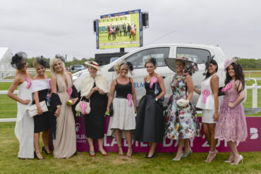 9 ladies dressed up for the horse races stood in front of a car and there is a TV screen with horces racing on it in the background.