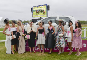 9 ladies dressed up for the horse races stood in front of a car and there is a TV screen with horces racing on it in the background.