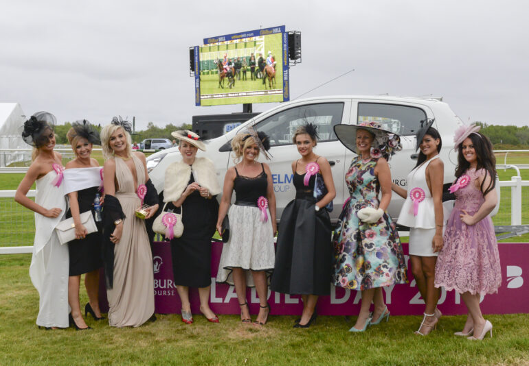 9 ladies dressed up for the horse races stood in front of a car and there is a TV screen with horces racing on it in the background.