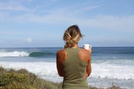 woman stood with her back to the camera looking out towards the ocean. She is holding a white coffee cop, her blonde hair is plaited to one sdie and she is wearing a khaki green racer back vest.