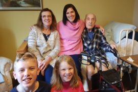 5 family members, 2 children a boy and a girl at the front and 3 adults two women and an elderly man sat on a bed at the back