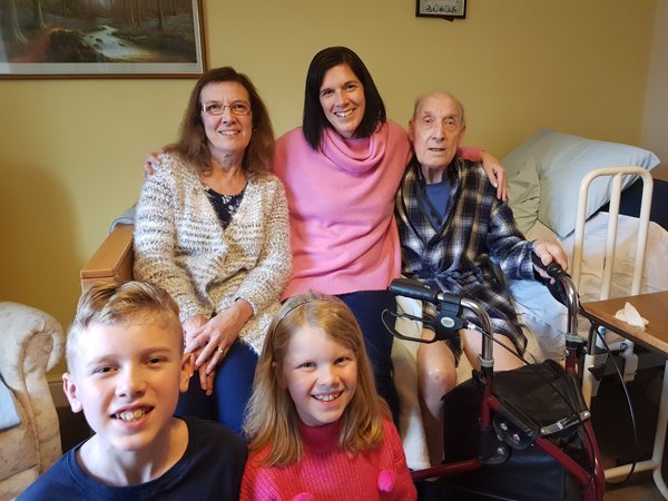 5 family members, 2 children a boy and a girl at the front and 3 adults two women and an elderly man sat on a bed at the back