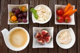 A selection of healthy foods including tomatoes, figs, hummus laid out in 6 white bowls on a wopoden surface