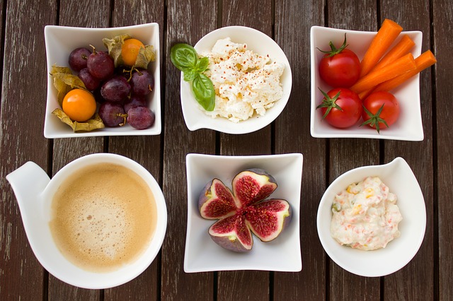 A selection of healthy foods including tomatoes, figs, hummus laid out in 6 white bowls on a wopoden surface