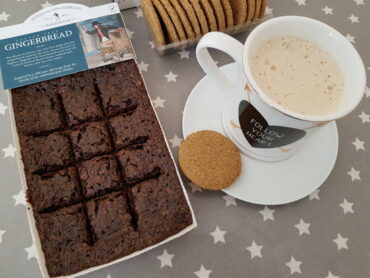 Horsham gingerbread in it's tray packet with a mug of cappucino on a plate and some biscuits