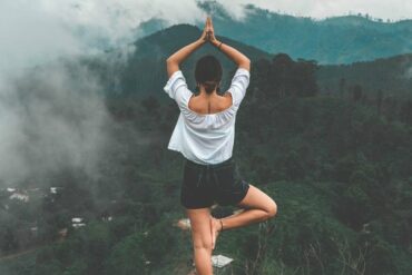 Woman stood on the edge of a mountain doing a yoga stand on one leg looking out across the jungle landscape