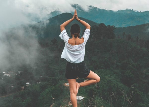 Woman stood on the edge of a mountain doing a yoga stand on one leg looking out across the jungle landscape