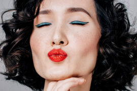 The face of a dark haired woman wearing lots of makeup = blue eye shadow, red lips, closing her eyes and pouting to the camera