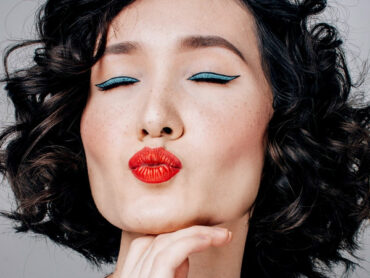 The face of a dark haired woman wearing lots of makeup = blue eye shadow, red lips, closing her eyes and pouting to the camera
