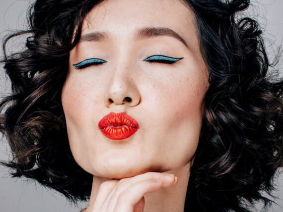 The face of a dark haired woman wearing lots of makeup = blue eye shadow, red lips, closing her eyes and pouting to the camera