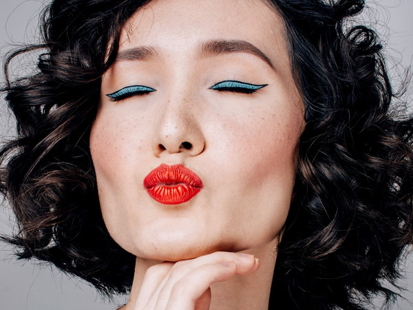 The face of a dark haired woman wearing lots of makeup = blue eye shadow, red lips, closing her eyes and pouting to the camera