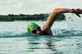 Man swimming in open water, he is wearing a green swimming hat and goggles