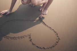 woman crouched down on a beach drawing a heart with her finger in the sand. you can only see her legs and her arms.