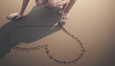 woman crouched down on a beach drawing a heart with her finger in the sand. you can only see her legs and her arms.