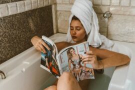woman lying in the bath with a big white towel on her head and she is reading a magazine having created a diy spa at home