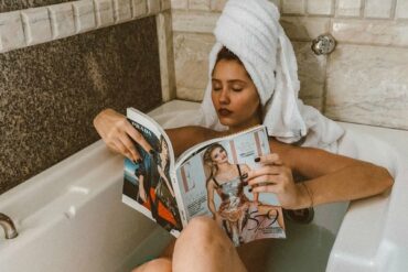 woman lying in the bath with a big white towel on her head and she is reading a magazine having created a diy spa at home