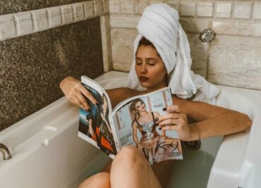 woman lying in the bath with a big white towel on her head and she is reading a magazine having created a diy spa at home