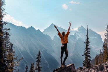 female stood on top of a rock wth arms outstretched. In the background s a range of mountains and the sky s blue with a few misty clouds.