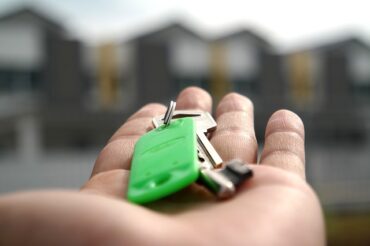 palm of someone's hand holdng a set of house keys on a green keyring in front of a blurry row of houses. It suggests they have just moved and got the keys to their first new home.