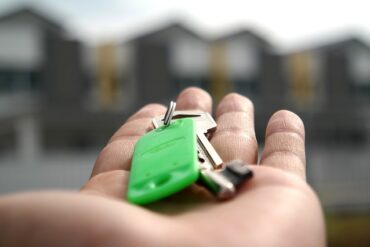 palm of someone's hand holdng a set of house keys on a green keyring in front of a blurry row of houses. It suggests they have just moved and got the keys to their first new home.