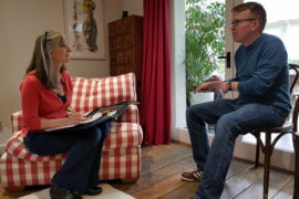 Annabel Boys the Body Stress Release practitioner is say n a red and white checked armchair on the left hand side of the picture. She has a folder on her lap and is askng questions to Simon Stafferton who is sat in a chair on the right. They are in her home studio and there is a pcture of the top half of the human skseleton on the wall.