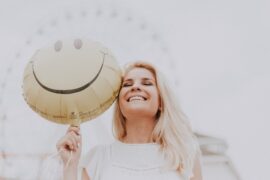 long blonde haired woman with a bright healthy smile holding a yellow smiley face helium balloon with a big wheel in the background