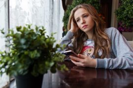 Teenage girl holding her mobile phone and staring sadly into the distance. She is sat ata a table with a plant on it and it struggling with cell phone addiction