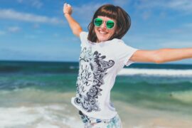 Young woman pictured from the waist up jumping into the air with joy. She is wearng sunglasses and a white t-shirt with print and is on a beack with the sea in the background.
