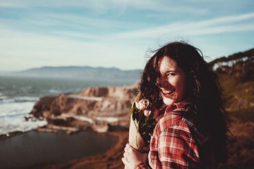 long haired brunette woman in her 40s stood overlooking mountains, she s laughing and looking healthy and care free. She is holdng a small bunch of brown paper wrapped pink roses.