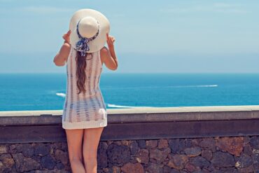 Female wearing a sun hat with a riboon and a white net sun dress that you can see her bikini through. She is stood to the left of the photo in front of a low wall overlooking the sea.
