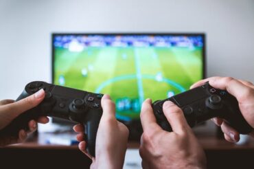 close up of two peoples hands holding game console controllers with a bg TV screen in th background that has a football video game on it.