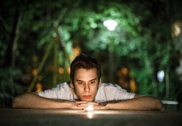 a young man resting his arms on a table looking into the flame of a candle. Going through the process of healing and learning he is not at fault.