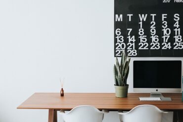Wooden office desk with two white chairs tucked under. There is a plant on the right hand sde and a reed diffuser on the left sde. On the wall there is half a black weekly calendar