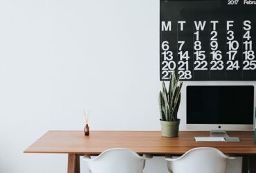 Wooden office desk with two white chairs tucked under. There is a plant on the right hand sde and a reed diffuser on the left sde. On the wall there is half a black weekly calendar