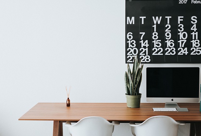 Wooden office desk with two white chairs tucked under. There is a plant on the right hand sde and a reed diffuser on the left sde. On the wall there is half a black weekly calendar