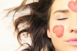 close up of half of a woman's face. Her eyes are closed and her lond brown hair is spread out to the side of her head on a white background. Shae has two red petals in the shape of hearts on her face, one on her cheek and one on her forehead. The images represents the health and wellness trends emerging for women in 2020