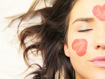 close up of half of a woman's face. Her eyes are closed and her lond brown hair is spread out to the side of her head on a white background. Shae has two red petals in the shape of hearts on her face, one on her cheek and one on her forehead. The images represents the health and wellness trends emerging for women in 2020